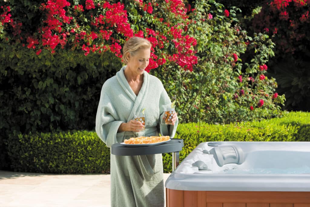 a woman in a robe uses hot tub accessories while standing next to her hot tub with flowers in the background