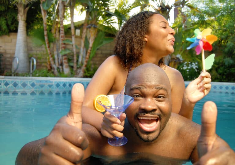 A man standing in a swimming pool is smiling at the camera giving the thumbs up sign with both hands. There is a woman behind him. She is resting her wrist on the man's shoulder and has a drink in that hand. She is holding a pinwheel with the other hand and is blowing towards it.