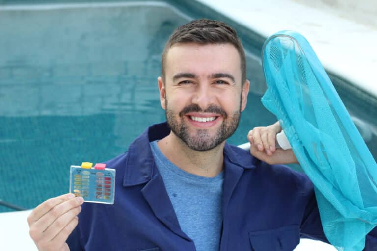 A man is holding a pool skimming net and a pool test kit. He is standing in front of a pool and smiling.
